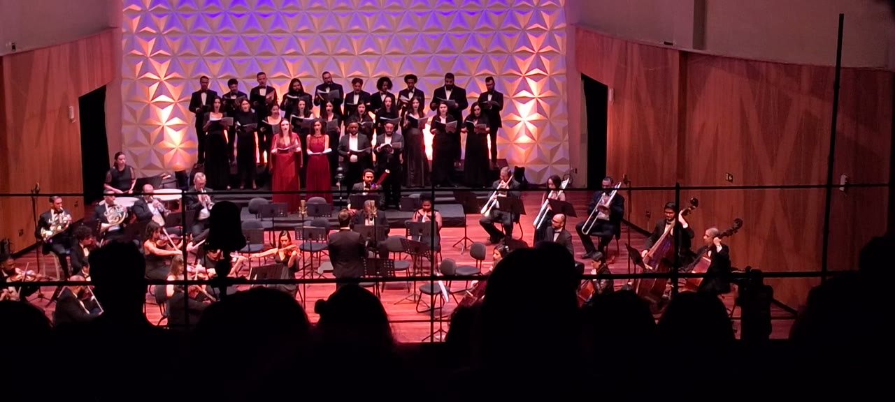 Coro e orquestra em concerto no palco de uma sala de concertos, com solistas ao centro